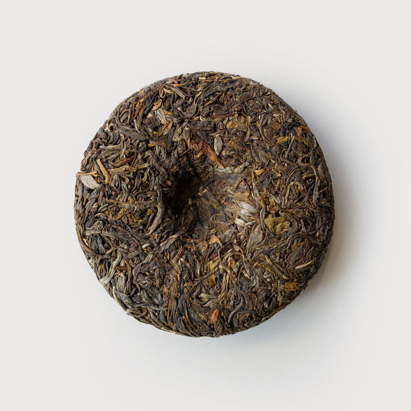 A Wa Shan Sheng Pu'er Cake Vintage 2022 by Rishi Tea & Botanicals on a white surface, surrounded by 200 cakes in a Wa ethnic village.