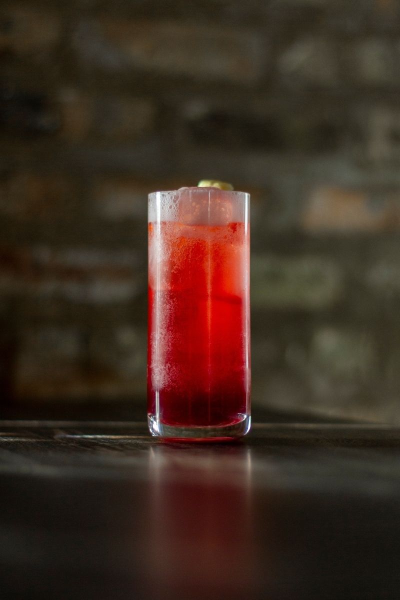 clear Collins glass filled with rich red cocktail in front of brick wall