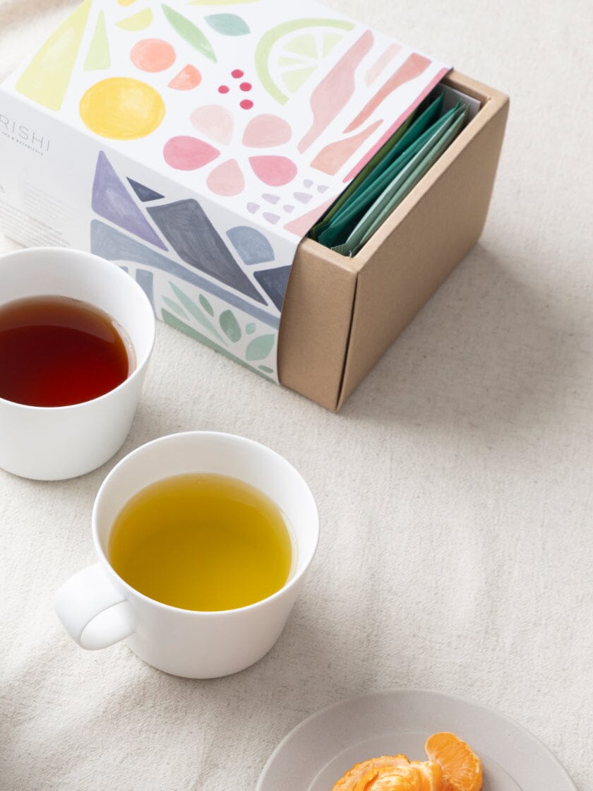 Tea bags in a colorful box with two cups of tea and an orange segment on a table.