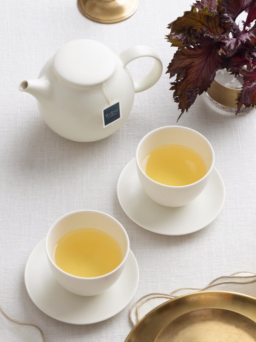 A white teapot with a tea bag label beside two filled teacups on saucers. A brass tray and red-purple leaves are visible on a white tablecloth.