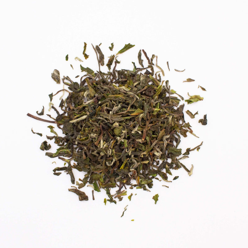A small pile of Darjeeling First Flush Chamong EX1 by Rishi Tea & Botanicals on a white background. The dried loose-leaf tea showcases green and brown hues, with subtle hints of stone fruits interwoven among the fragrant blossoms.