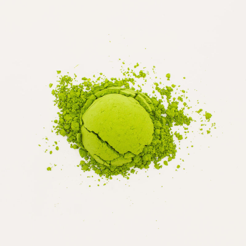 A crumbled macaron infused with Nishi San's Matcha Asatsuyu by Rishi Tea & Botanicals lies on a white background, its fragments scattered artfully around.