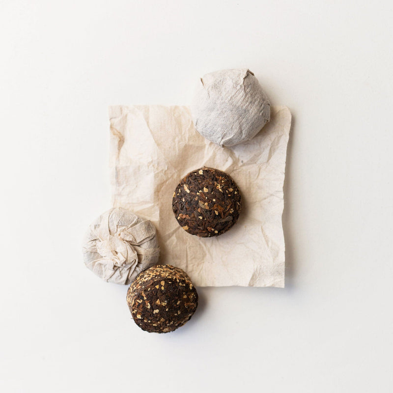 Four wrapped Pu'er Tuo Cha Cinnamon Ginger cakes from Rishi Tea & Botanicals sit on crumpled parchment, while two unwrapped dark cakes reveal textured surfaces and an invigorating aroma.