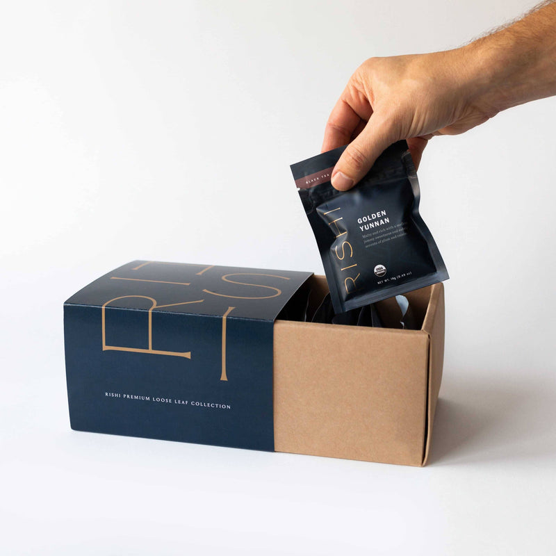 A hand holding a small black packet of the Premium Loose Leaf Tea Collection from Rishi Tea & Botanicals above an open cardboard box filled with similar packets.