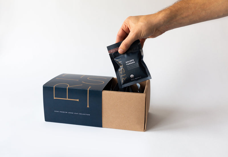 A hand holding a small black packet of the Premium Loose Leaf Tea Collection from Rishi Tea & Botanicals above an open cardboard box filled with similar packets.