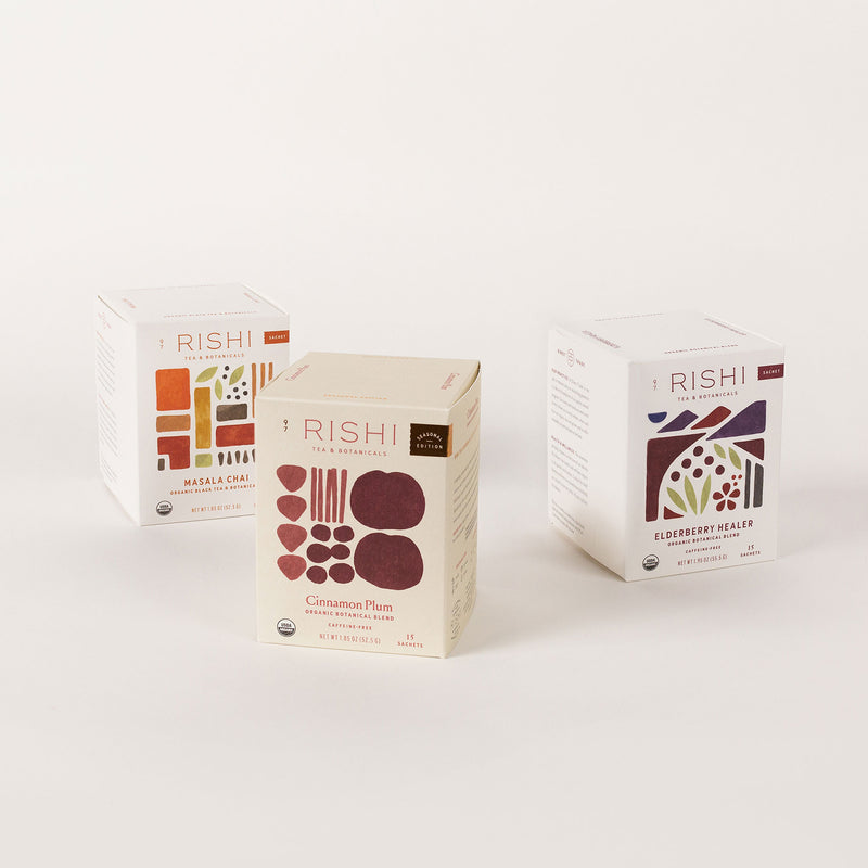 Displayed are three boxes from the Winter Sachet Collection by Rishi Tea & Botanicals: Cinnamon Plum, Blueberry Hibiscus, and one with a blurred label. Each box showcases colorful geometric designs that capture the cozy essence of winter-inspired sachet teas.