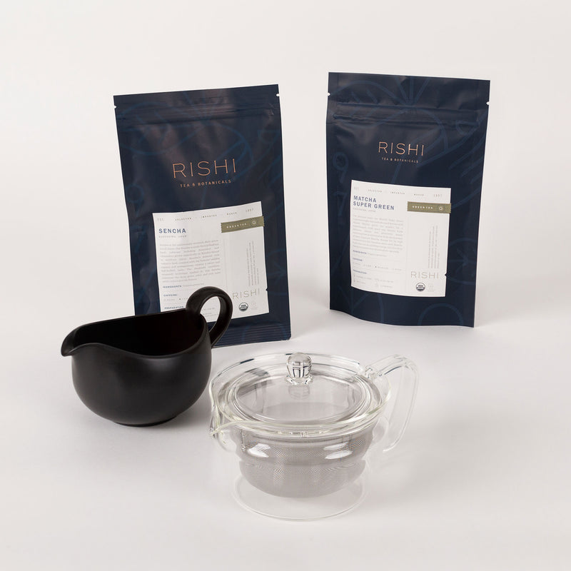 The Japanese Green Tea Starter Bundle from Rishi Tea & Botanicals, including Matcha Super Green and Sencha packages, a black pitcher, and a clear teapot with an infuser, is displayed on a plain background.