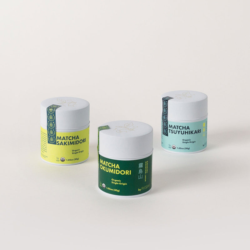 The Single-Origin Matcha Trio Bundle from Rishi Tea & Botanicals features three cylindrical containers of organic ceremonial matcha named Sakimidori, Okumidori, and Tsuyuhikari. Each container is capped with a white lid and set against a light background, offering unique single-cultivar flavors for an exquisite tea experience.